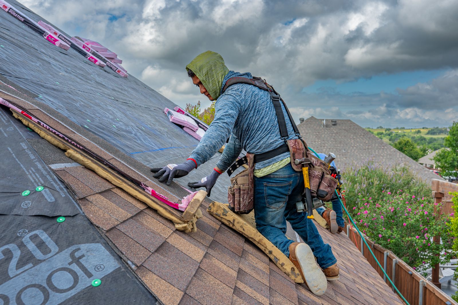 Harnessed roofer installing asphalt shingles above a suburban neighborhood